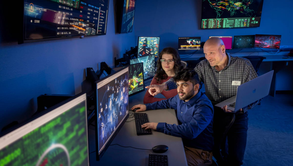 Three people are in a dimly lit room with multiple computer screens displaying data. A man with a laptop stands, another man seated is pointing at one screen, and a woman sits beside them