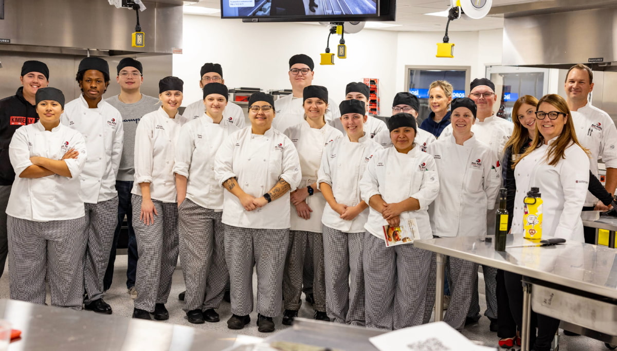 A group of chefs and culinary students pose in a kitchen. They are wearing white chef coats and black hats. The kitchen is equipped with stainless steel counters and cooking equipment. Everyone is smiling and looking at the camera