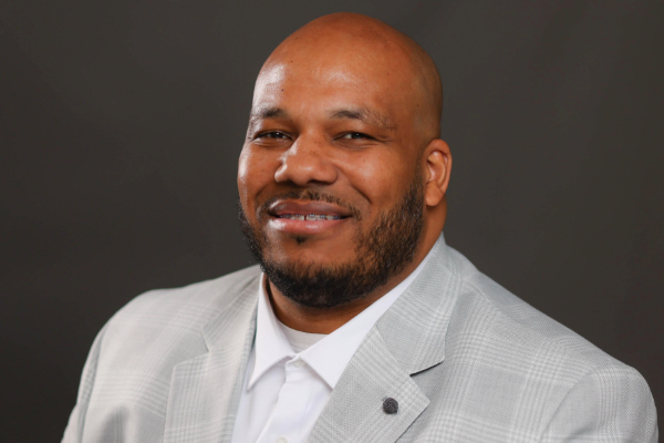 A man with a beard wearing a light gray plaid suit and a white shirt, smiling at the camera with a neutral background