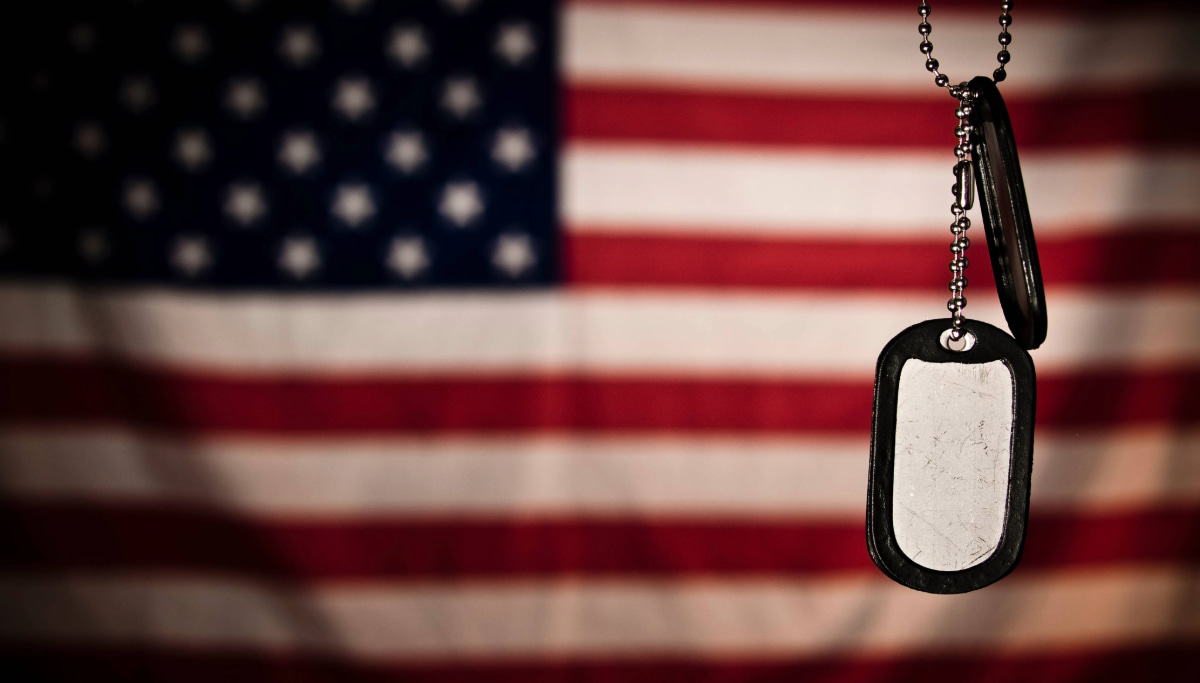 Dog tags on a chain are suspended in front of a blurred background of the American flag