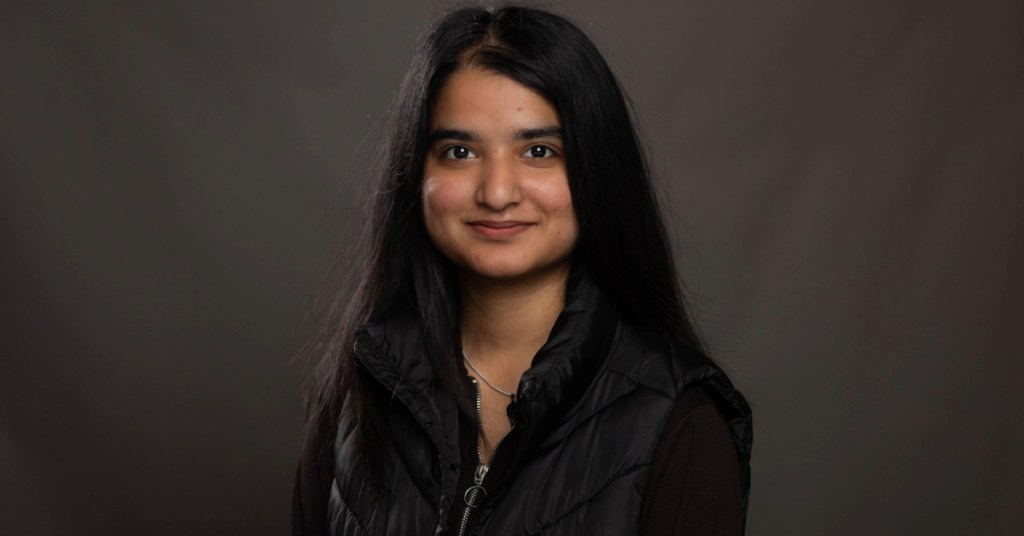 A person with long, dark hair smiles slightly while wearing a black puffy vest over a dark top. The background is a simple, neutral gray, giving a professional portrait appearance