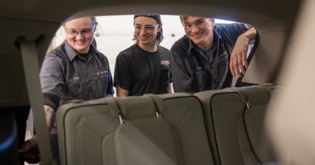 Three people in work uniforms and safety glasses smiling and examining the interior of a vehicle from the rear