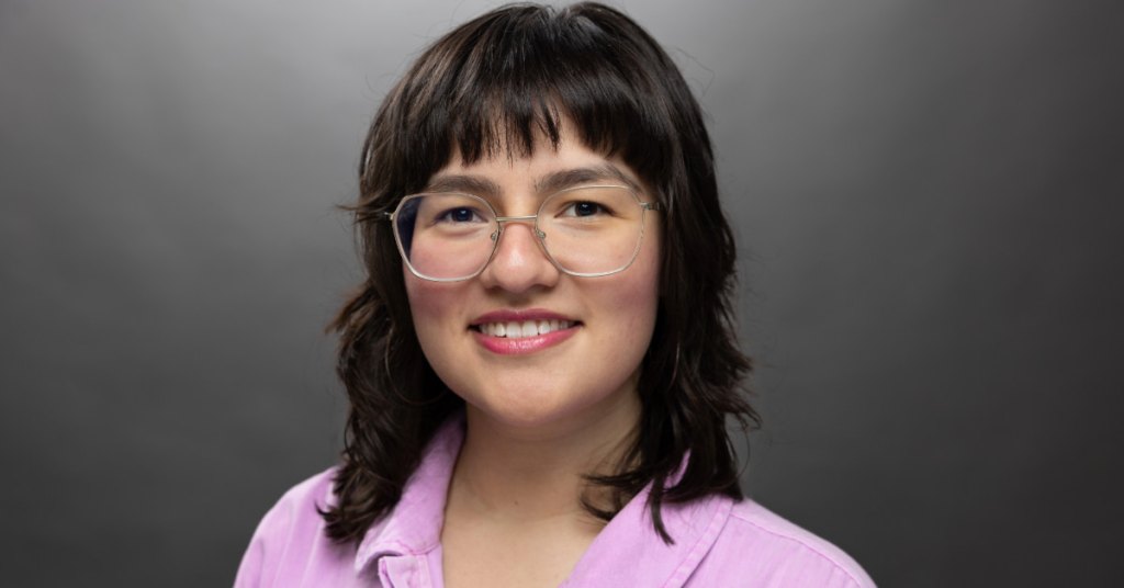 Person with shoulder-length dark hair, wearing glasses and a purple shirt, smiling against a gray background