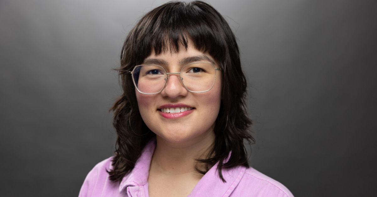 Person with shoulder-length dark hair, wearing glasses and a purple shirt, smiling against a gray background