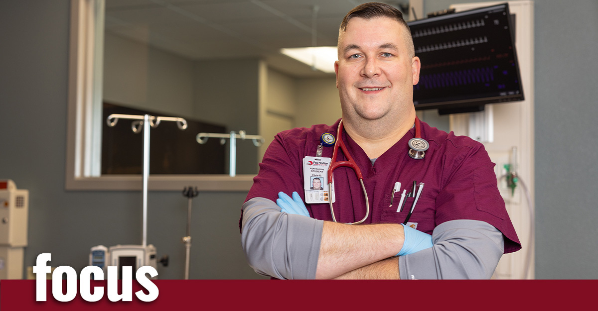 A healthcare professional in maroon scrubs, wearing a stethoscope, stands in a medical room