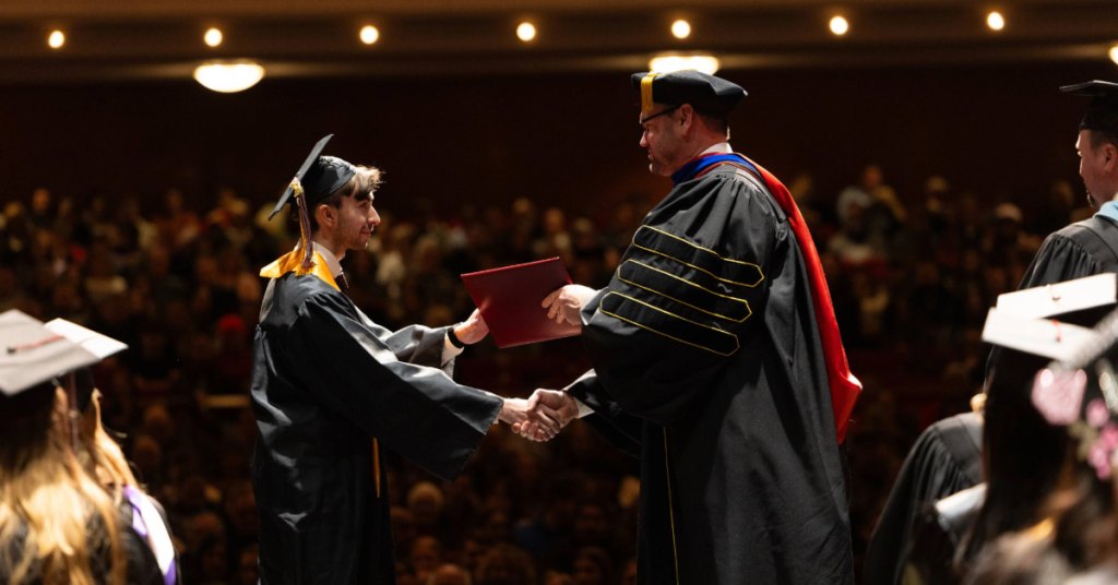 A graduate in a cap and gown shakes hands with FVTC president in academic regalia, receiving a diploma on stage. The background shows an audience seated in a dimly lit auditorium