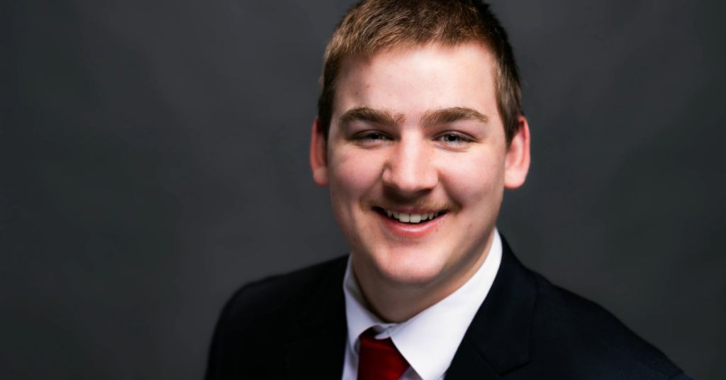 A person in a suit with a red tie smiles against a dark background