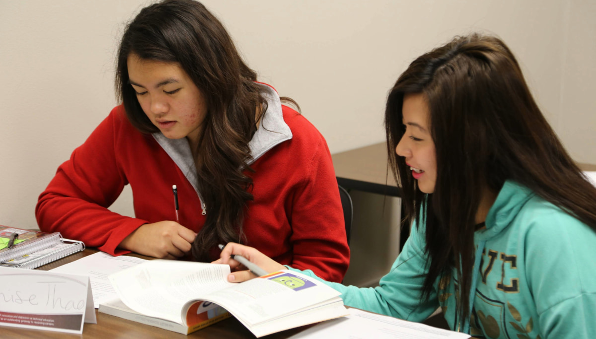 Two students are sitting at a table studying. One wears a red sweater and looks at a book, while the other, in a turquoise hoodie, points at the page