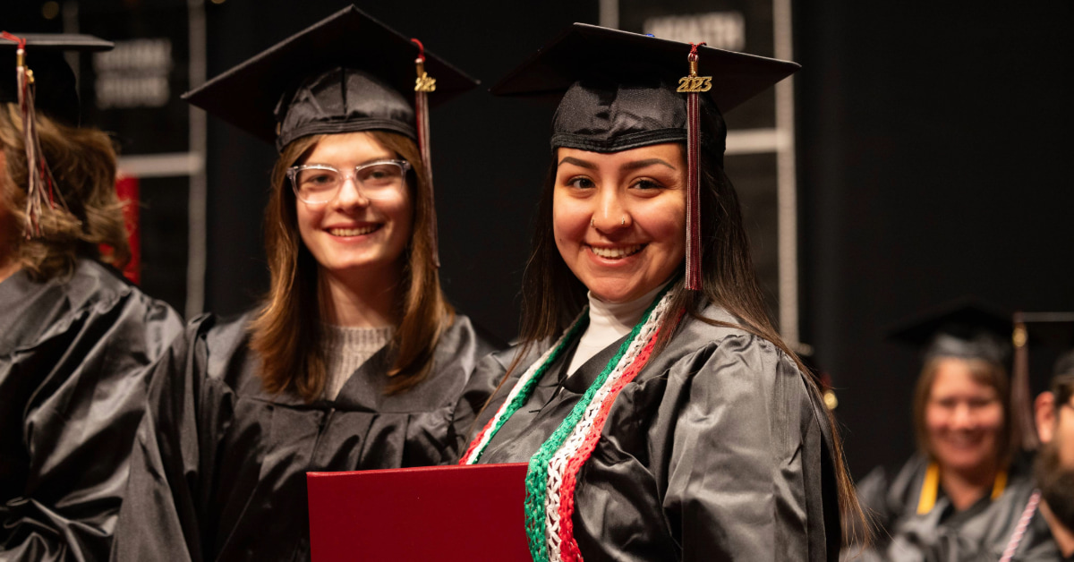 Two graduates in black caps and gowns smile at the camera. One holds a diploma folder and wears colorful cords