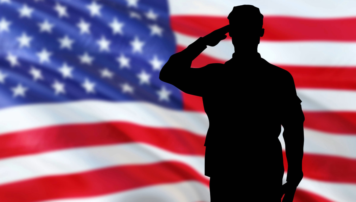 Silhouette of a person in military uniform saluting in front of a large, blurred American flag.
