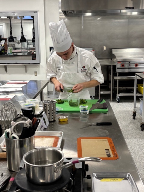 A culinary student in a kitchen prepares ingredients on a green cutting board, surrounded by cooking tools and containers.