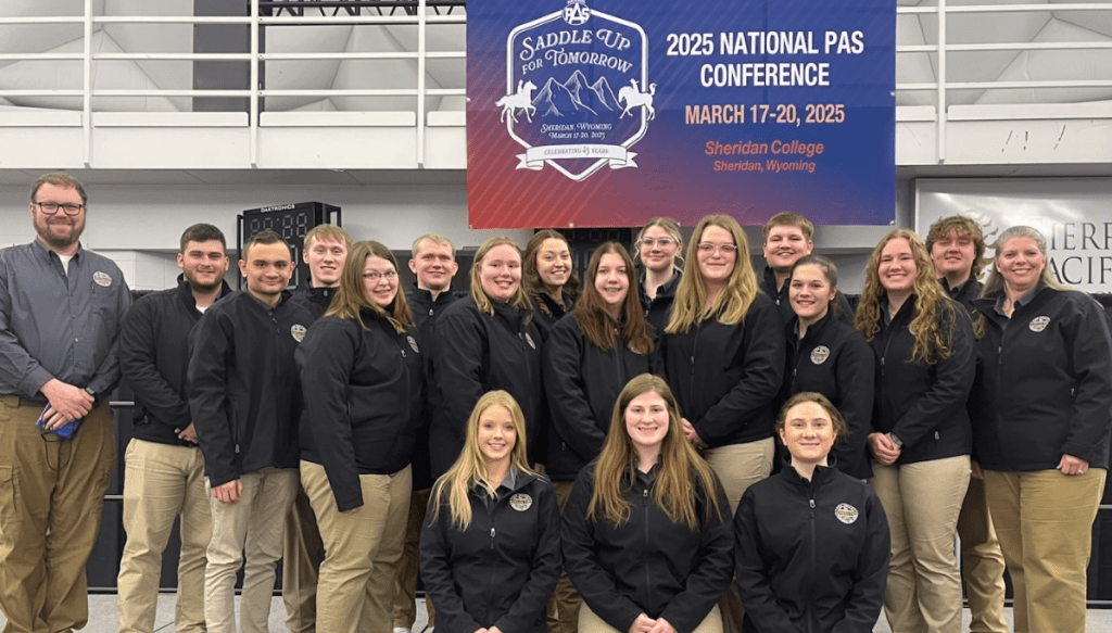 A group of people in matching jackets poses in front of a "2025 National PAS Conference" banner at Sheridan College.