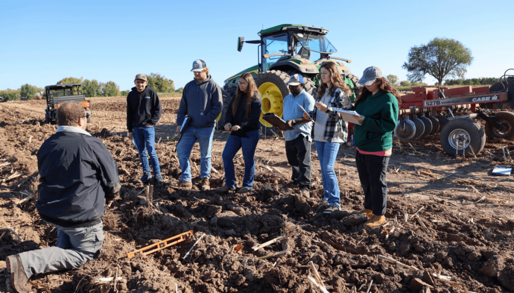 A group of people attentively listening to an instructor in a farm field, with a tractor and farming equipment in the background under a clear blue sky.