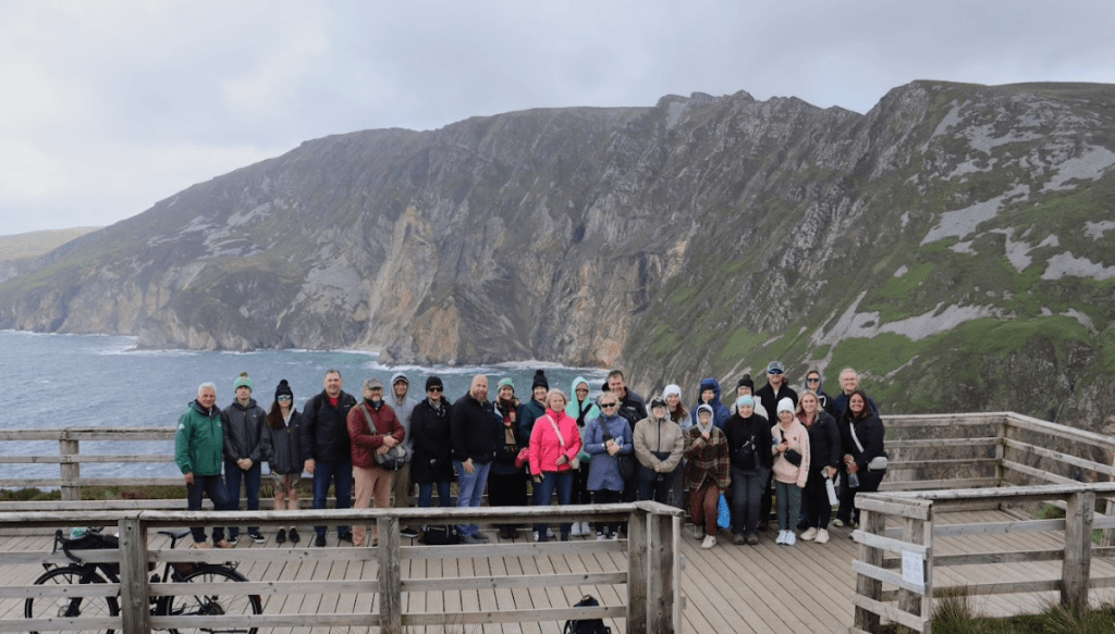 A group of people pose together on a wooden viewing platform overlooking dramatic coastal cliffs and the sea, with rugged green hills in the background under a cloudy sky.