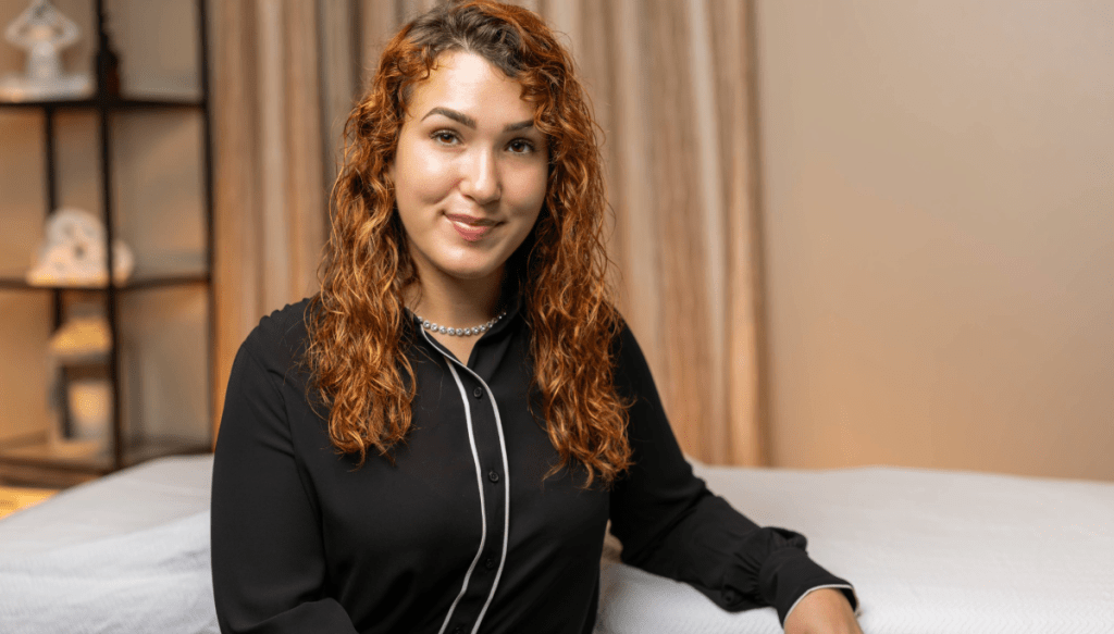 A woman with curly brown hair, wearing a black blouse, sits and smiles next to a massage table in a softly lit room with beige curtains and shelves in the background.