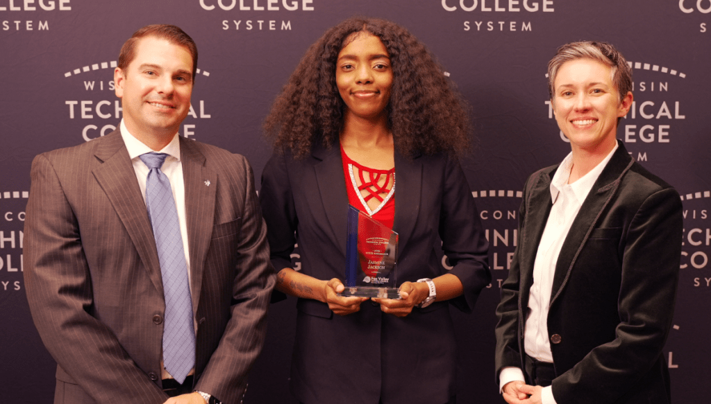 Three individuals smiling at an awards ceremony, with the central person holding a trophy. They are standing in front of a backdrop that reads "Wisconsin Technical College System.