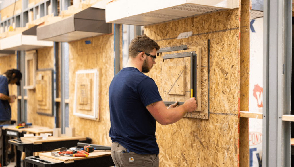 A man wearing glasses measures a window frame on a plywood wall in a workshop, with tools and materials on a nearby workbench. Another person works in the background.