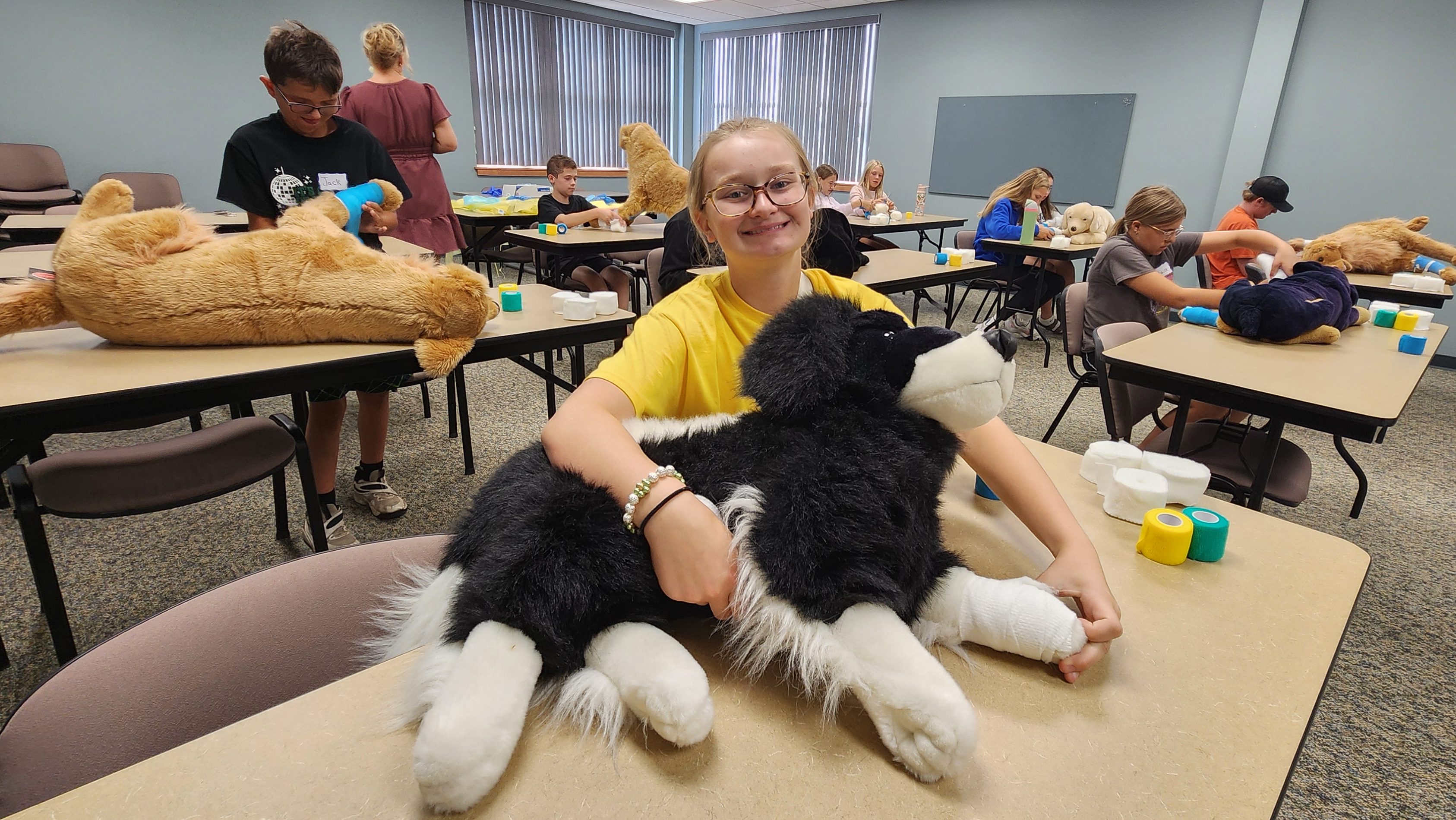A smiling girl is in the foreground of a room with children examining stuffed animals