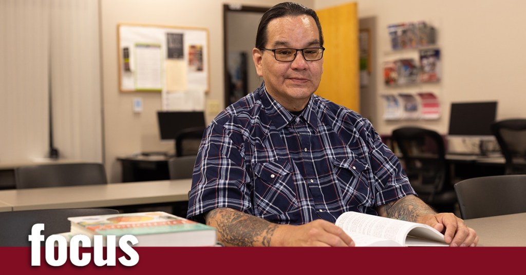 A man with tattoos sits at a desk in a classroom with books in front of him
