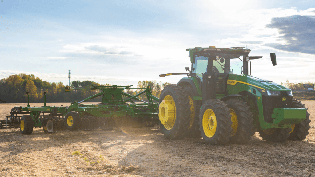 A green and yellow tractor with large tires pulls a disc harrow across a sunlit, harvested field, with trees and a partly cloudy sky in the background.