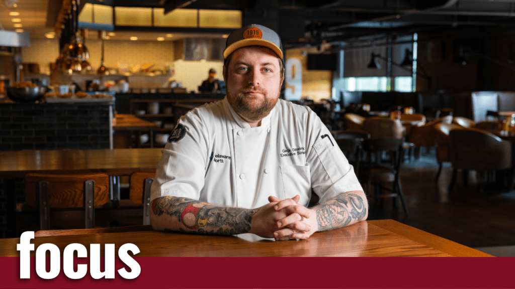 A chef with tattoos on both arms sits at a wooden table in a restaurant, wearing a white chefs coat and an orange cap. The background shows a modern, well-lit dining area and an open kitchen.