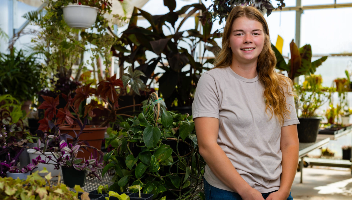 A young woman with long, light brown hair and a beige t-shirt stands smiling in a greenhouse filled with various green and purple potted plants. Sunlight streams through the windows behind her.