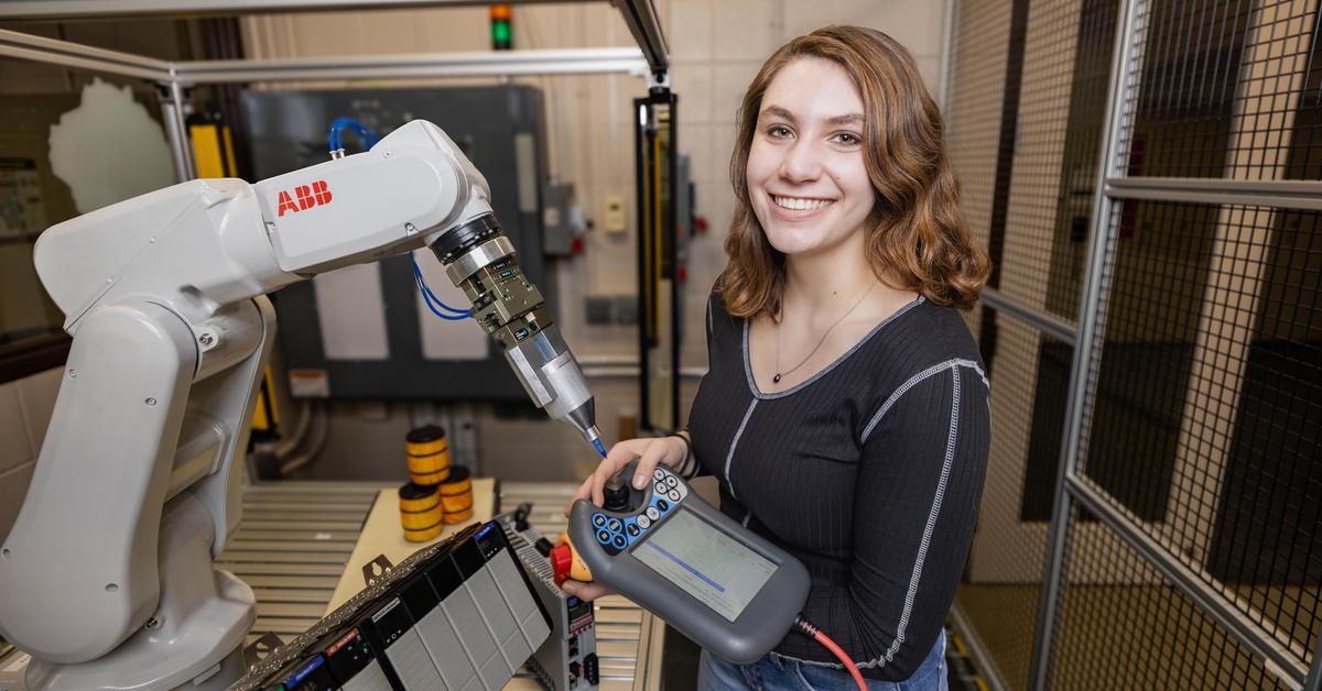 Katie Karow smiles while holding a controller, standing near a robotic arm.