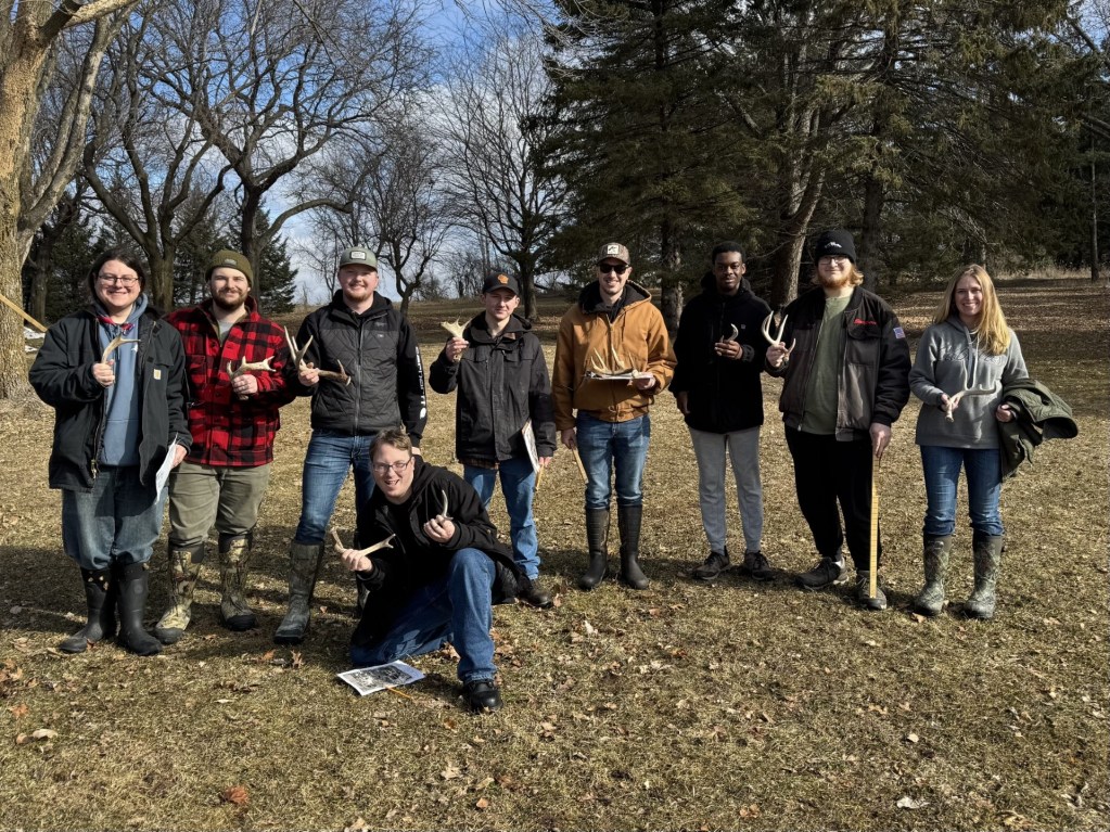 Nine people stand and kneel outdoors on grass, holding deer antlers and papers, dressed in jackets and boots. Leafless trees and a blue sky are in the background, suggesting early spring or late fall.