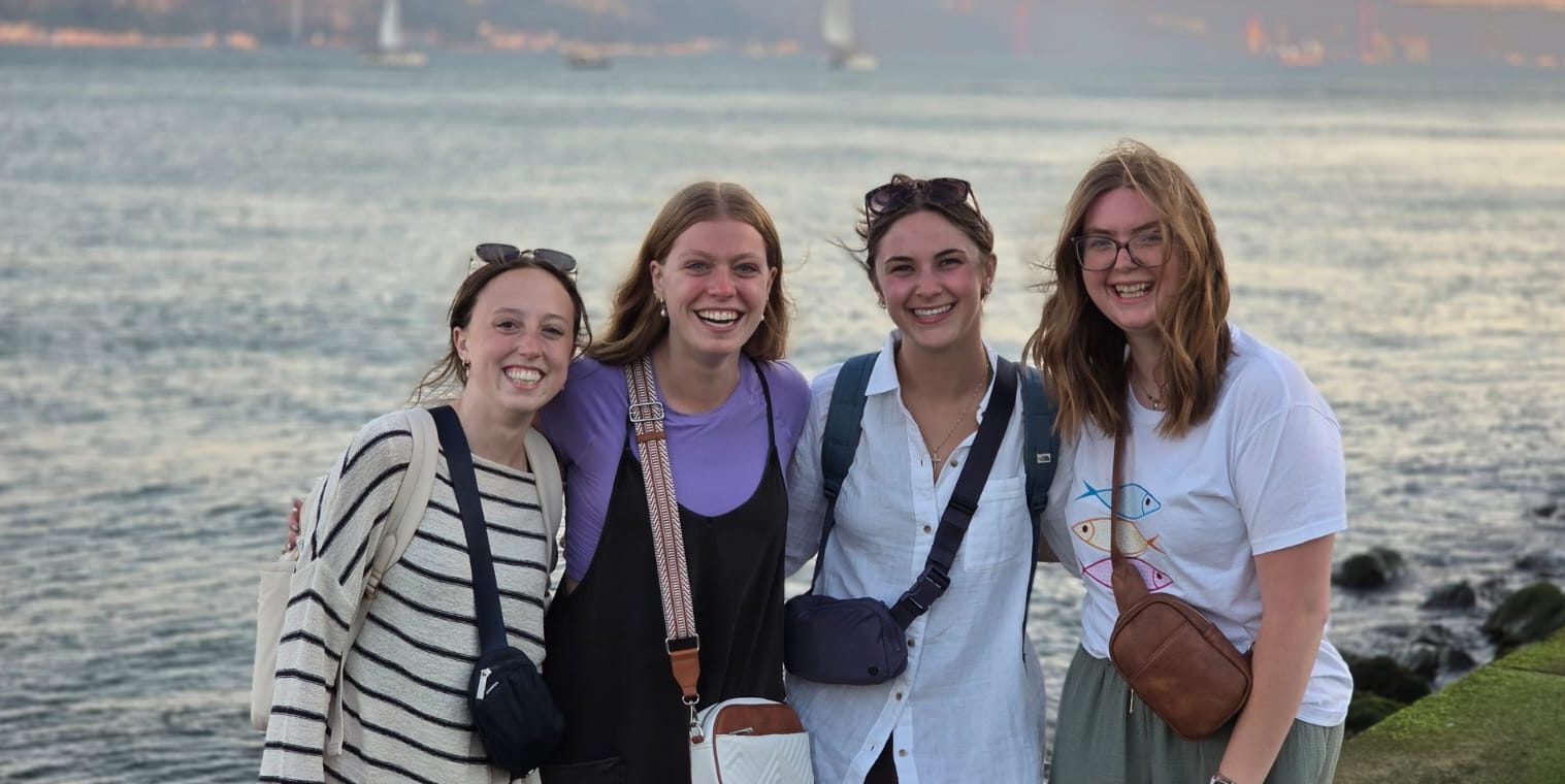 Four girls stand smiling in front of a body of water.