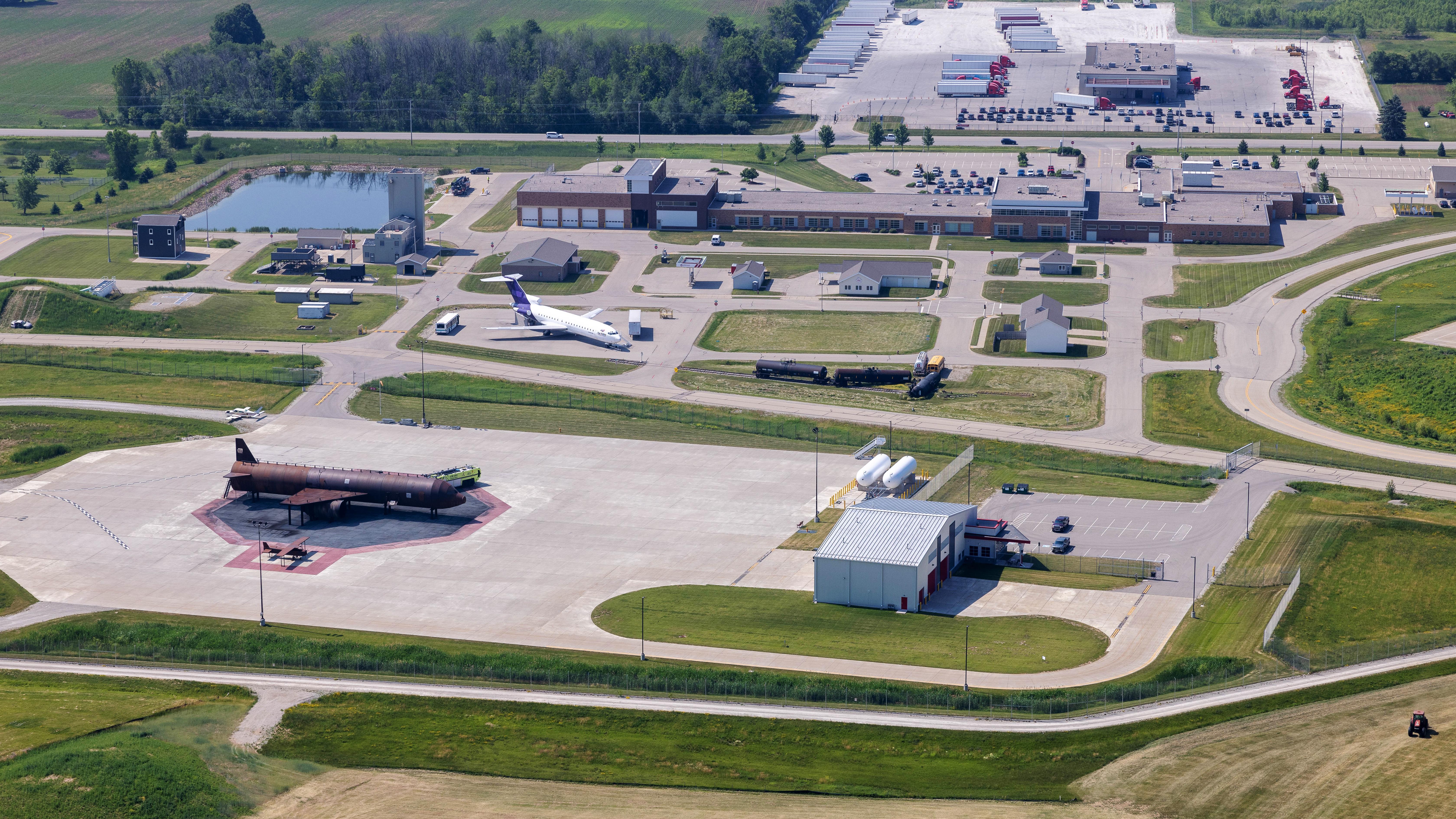Aerial view of an industrial complex with a white airplane, a large dark aircraft, a pond, and numerous trucks and buildings.