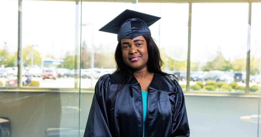 Nadege Jolivard stands against a glass wall, wearing a cap and gown over a bright blue top.