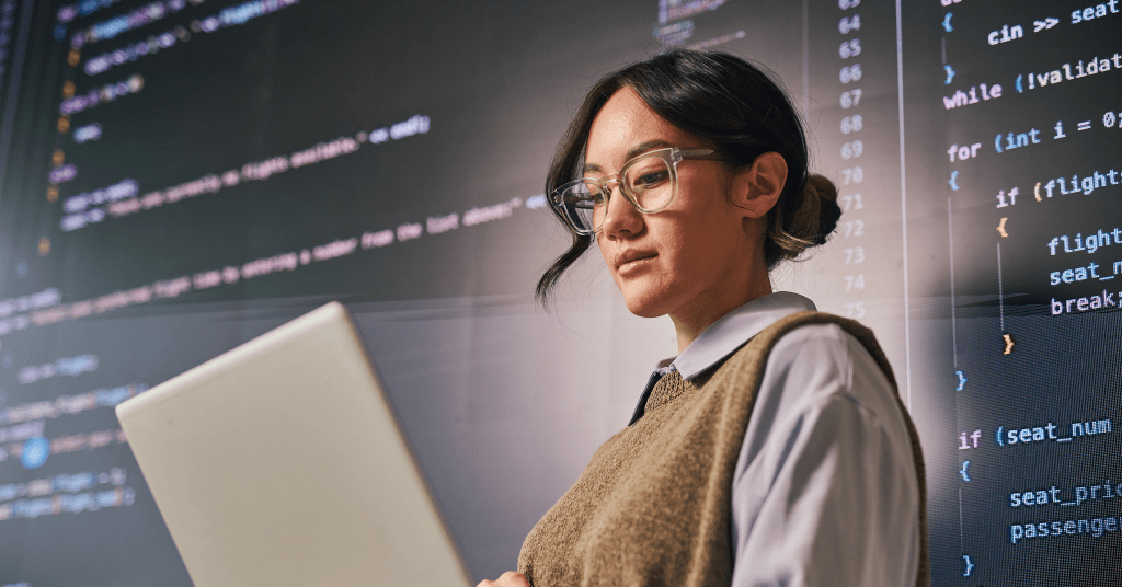 Woman wearing glasses and a sweater vest looks at a laptop, surrounding by code in the background.