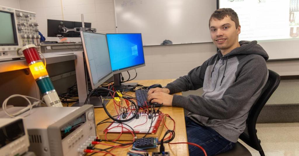 Male student in a gray hoodie sits at a computer. He is surrounded by black and red wires connected to machines, and a traffic light apparatus.