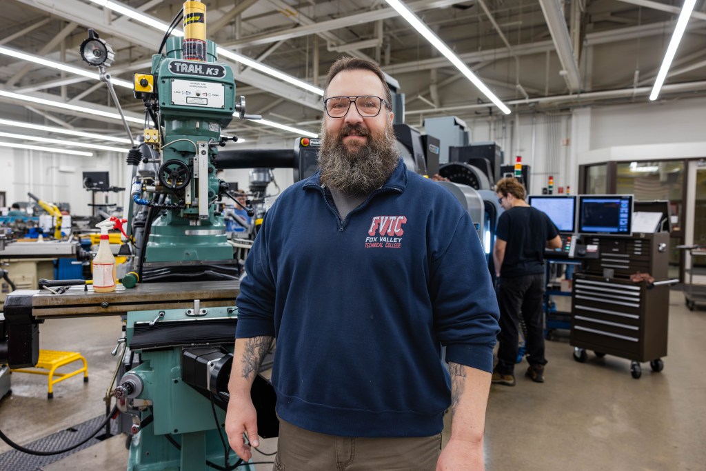 A man wearing glasses and a navy FVTC Fox Valley Tech shirt stands in a workshop with machinery and another person working in the background.