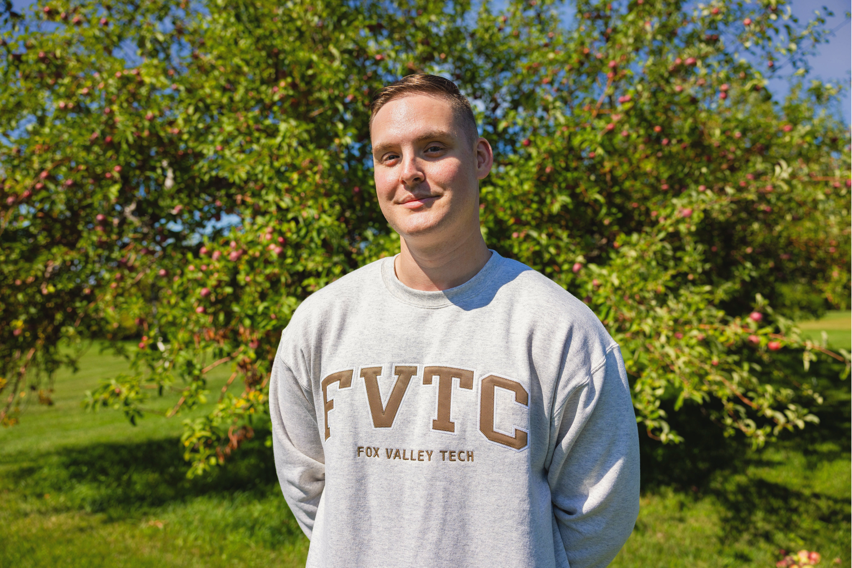 Alex Soliday stands in front of a large apple tree while wearing a brown FVTC sweatshirt.